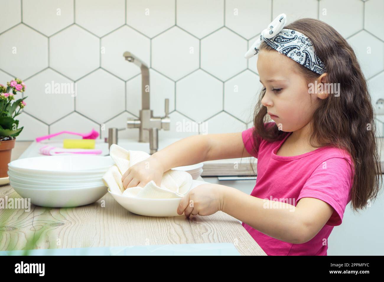 Contrarié petite fille essuyant des bols transparents avec une serviette sèche sur le set de cuisine. Portrait de l'enfant faisant la vaisselle dans l'évier de cuisine. Banque D'Images