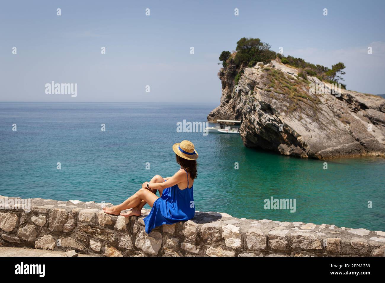 La fille dans une robe bleue et un chapeau de paille seule est assise près de la mer sur un rocher, sur une falaise et regarde la mer. Vacances, voyages, tourisme. Banque D'Images