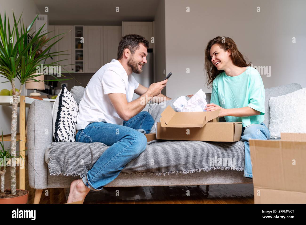 Jeune couple marié assis sur un canapé à la maison et déballant des boîtes en carton avec leur achat en ligne. Banque D'Images Jeune couple marié assis sur un canapé à la maison et déballant des boîtes en carton avec leur achat en ligne. Banque D'Images
