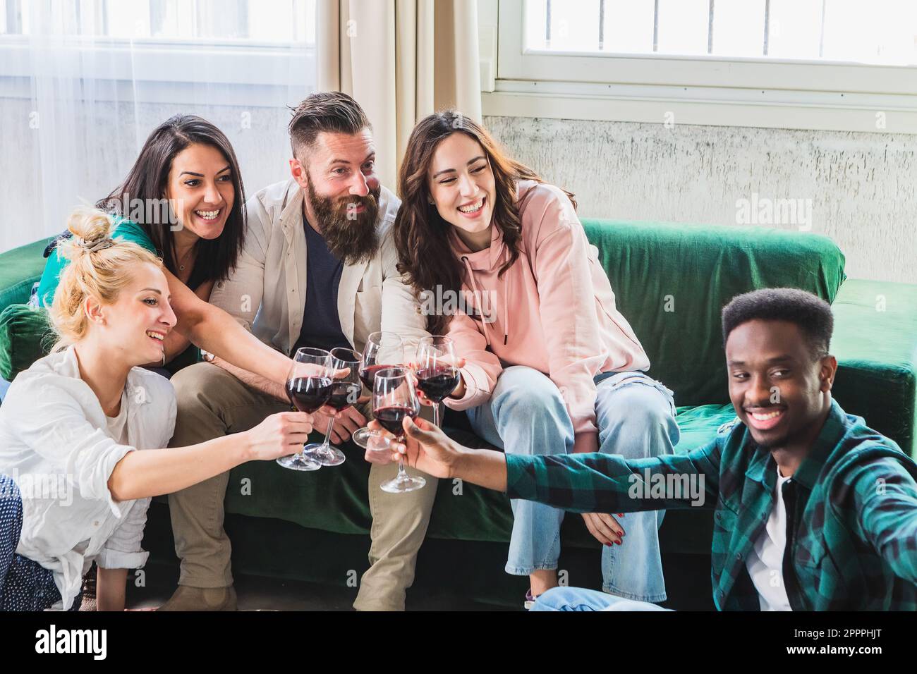 Groupe d'amis pour un apéritif à la maison avec vin rouge assis sur le canapé. Les personnes multiethniques du millénaire apprécient la fête qui se réunit. La vie Banque D'Images