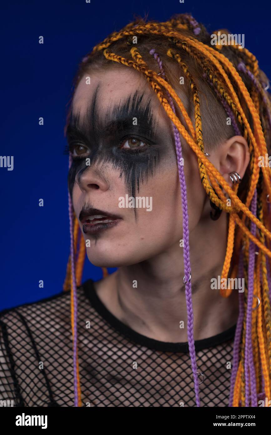 Jeune femme avec des tresses de couleur orange cheveux et scène noire créative maquillage peint sur le visage. Studio tourné sur fond bleu. Fait partie d'une série de photos Banque D'Images