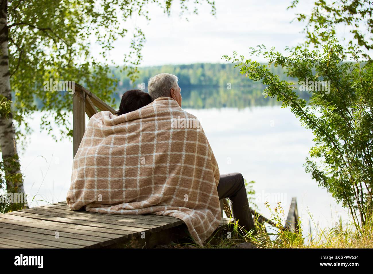 Couple d'âge mûr assis sur des marches en bois et embrassant. Ils sont enveloppés dans une couverture, souriant et regardant la vue magnifique - forêt et lac. Joyeux senior Banque D'Images
