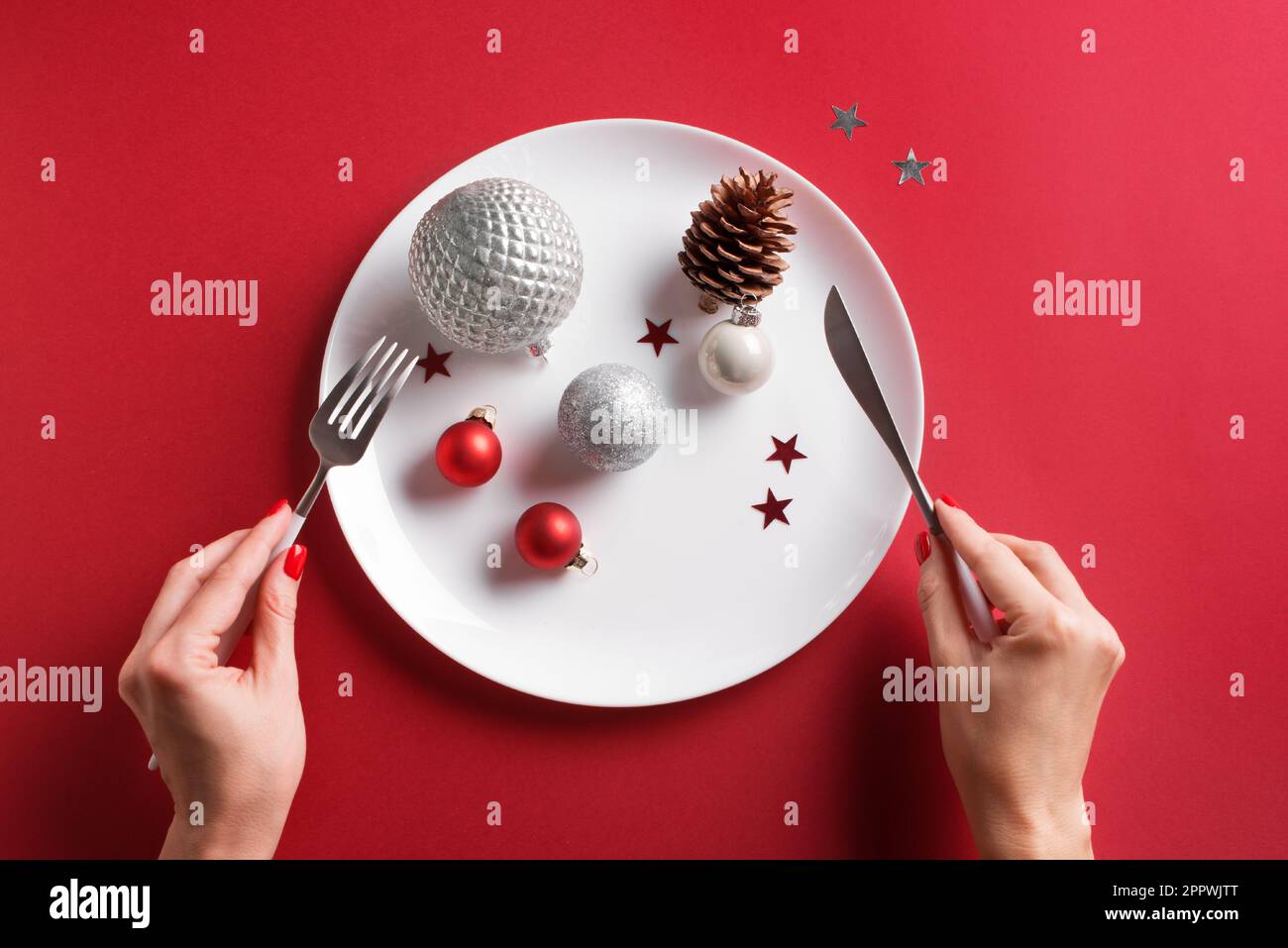 Magnifique table de Noël et mains de femmes. Boules argentées festives et cône de pin sur plaque blanche sur fond rouge, pose à plat, espace de copie. EATI Banque D'Images