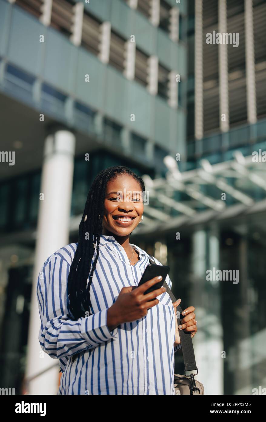 Bonne femme d'affaires noire souriant à l'appareil photo sur le chemin du travail en ville. Portrait d'une jeune femme d'affaires joyeuse utilisant un smartphone pendant que c Banque D'Images