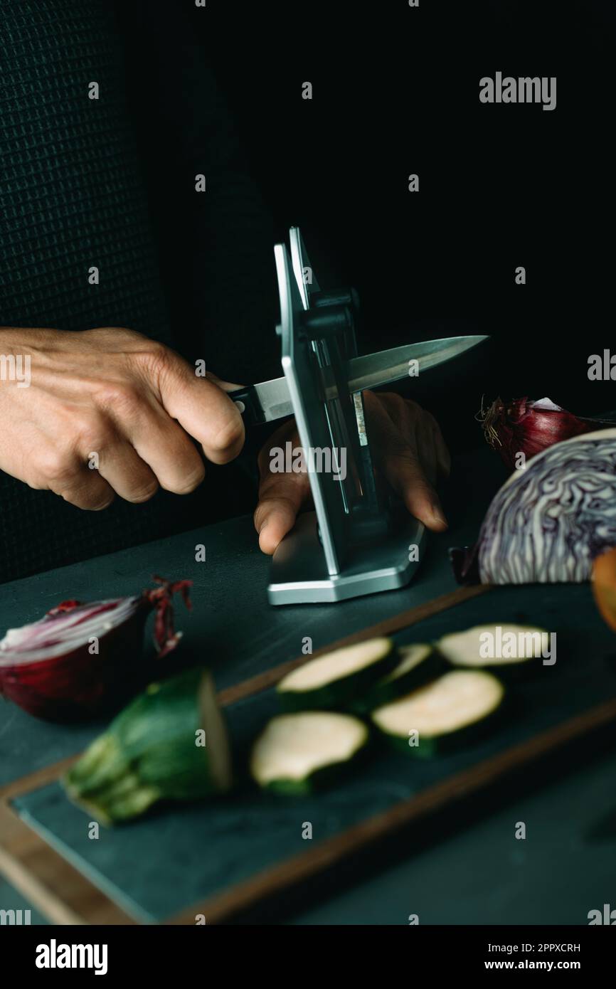 un homme aiguise un couteau de cuisine à l'aide d'un taille-crayon de bureau sur une table avec des légumes coupés et hachés Banque D'Images