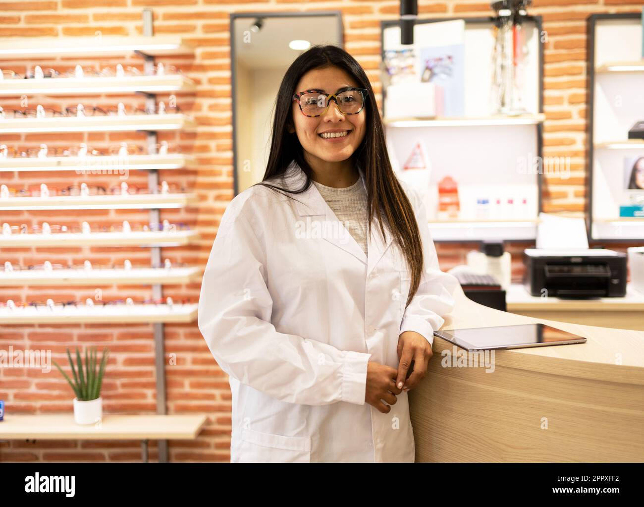 Un jeune ophtalmologiste positif avec des lunettes optiques debout près de la table avec une tablette et regardant la caméra tout en travaillant dans la boutique optique Banque D'Images