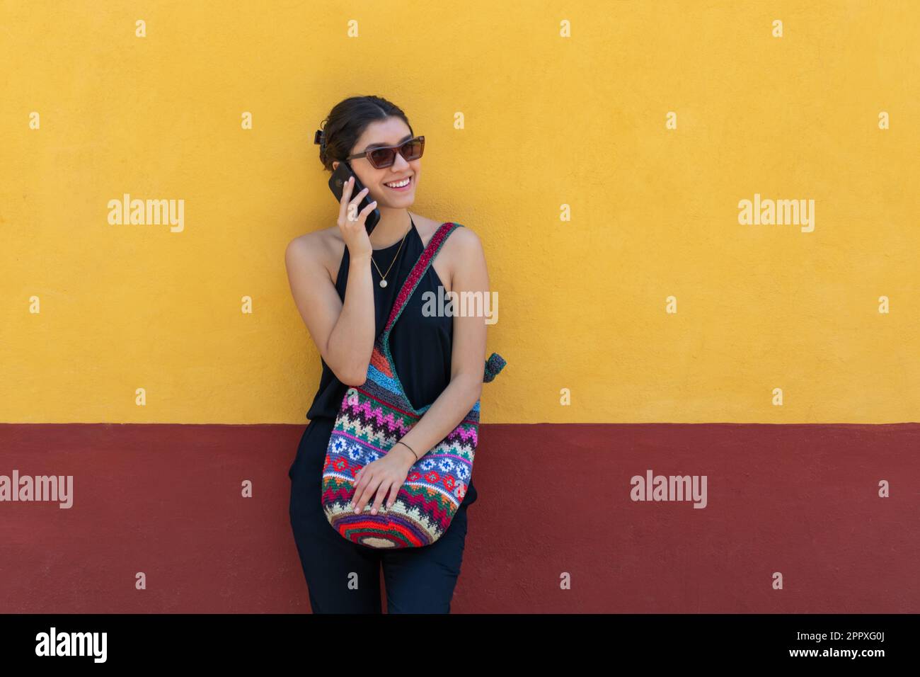 Jeune femme positive dans une tenue tendance et des lunettes de soleil souriant et regardant loin tout en se tenant contre le mur jaune et parlant sur le smartphone à l'extérieur dans Banque D'Images