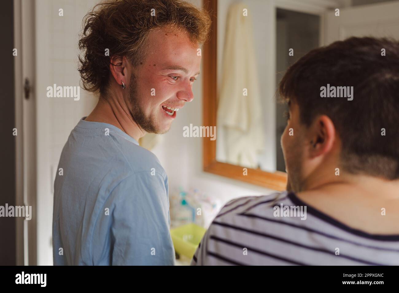 Des jeunes amis masculins positifs dans des t-shirts domestiques souriant et riant tout en regardant les uns les autres dans la salle de bains pendant la routine quotidienne Banque D'Images Des jeunes amis masculins positifs dans des t-shirts domestiques souriant et riant tout en regardant les uns les autres dans la salle de bains pendant la routine quotidienne Banque D'Images