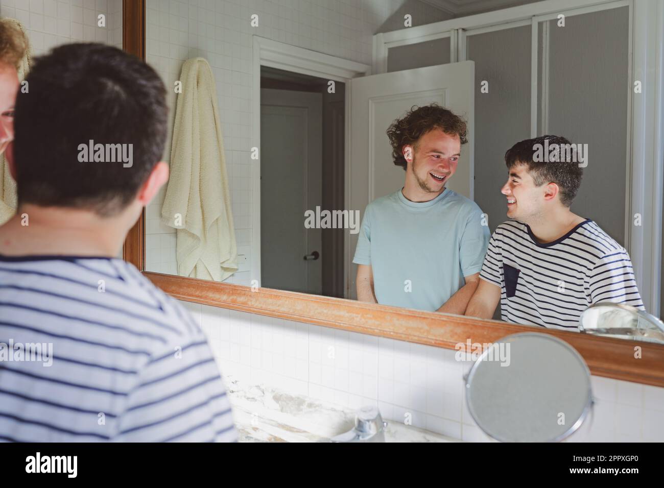 Des jeunes amis masculins positifs dans des t-shirts domestiques souriant et riant tout en regardant les uns les autres dans la salle de bains pendant la routine quotidienne Banque D'Images Des jeunes amis masculins positifs dans des t-shirts domestiques souriant et riant tout en regardant les uns les autres dans la salle de bains pendant la routine quotidienne Banque D'Images