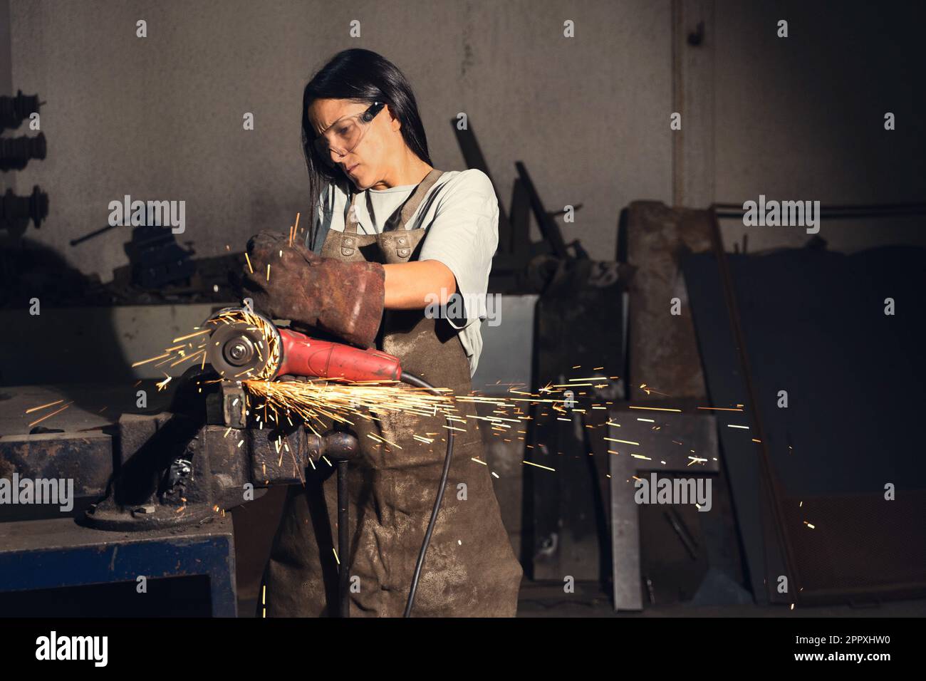 Jeune femme concentrée dans un tablier de forge et des gants portant des lunettes de protection et de coupe de détail de fer avec meuleuse d'angle dans les étincelles volantes pendant que W Banque D'Images