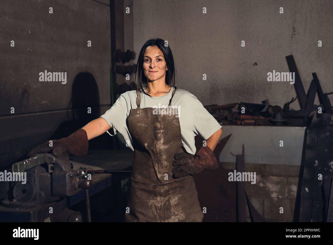 Jeune femme forgeron positif en uniforme et gants souriant et regardant la caméra tout en travaillant avec l'équipement professionnel pendant le travail Banque D'Images