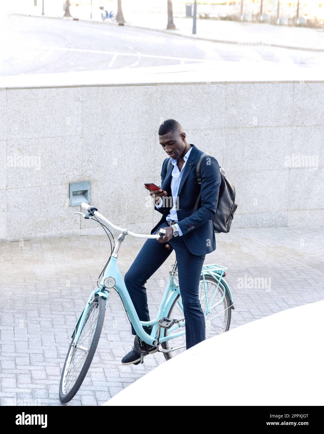 Un homme d'affaires afro-américain sérieux portant un costume habillé assis sur un vélo et parcourant un téléphone portable tout en parcourant une rue près d'un mur en béton Banque D'Images