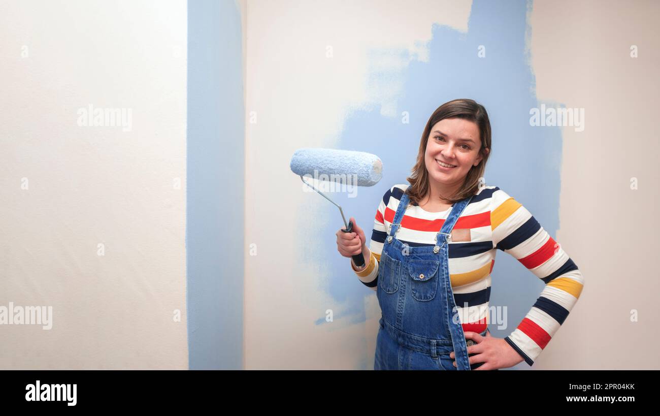 Belle femme debout vêtue de combinaisons, souriant à l'appareil photo avec rouleau de peinture bleu dans sa main sur fond de mur blanc à moitié peint Banque D'Images