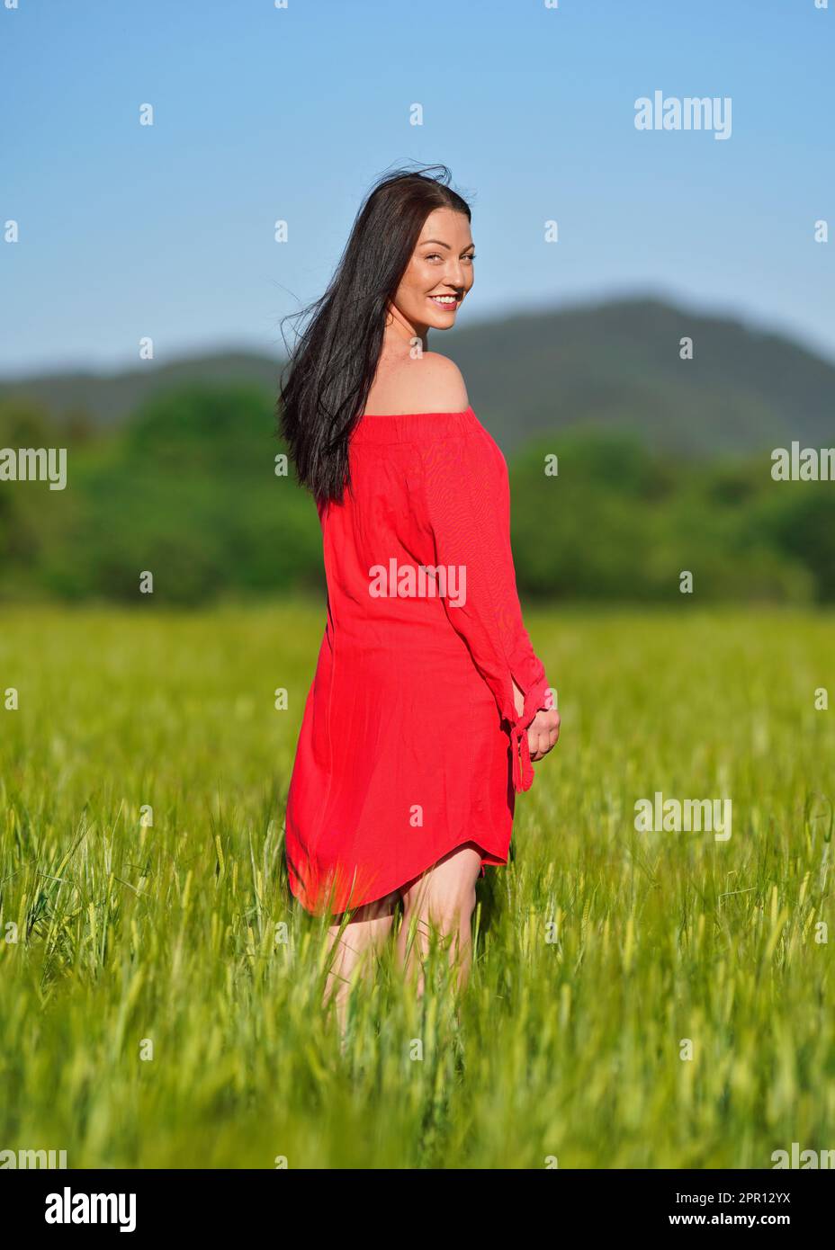Jeune femme en robe rouge d'été marchant dans le champ vert de blé non mûr, regardant en arrière par-dessus son épaule Banque D'Images