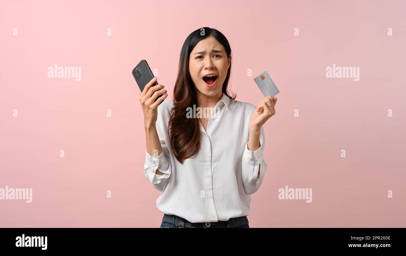 Choqué et surpris jeune femme asiatique tenant son smartphone et sa carte de crédit, isolée sur fond rose. nouvelle promotion, offre spéciale, achats en ligne. Banque D'Images
