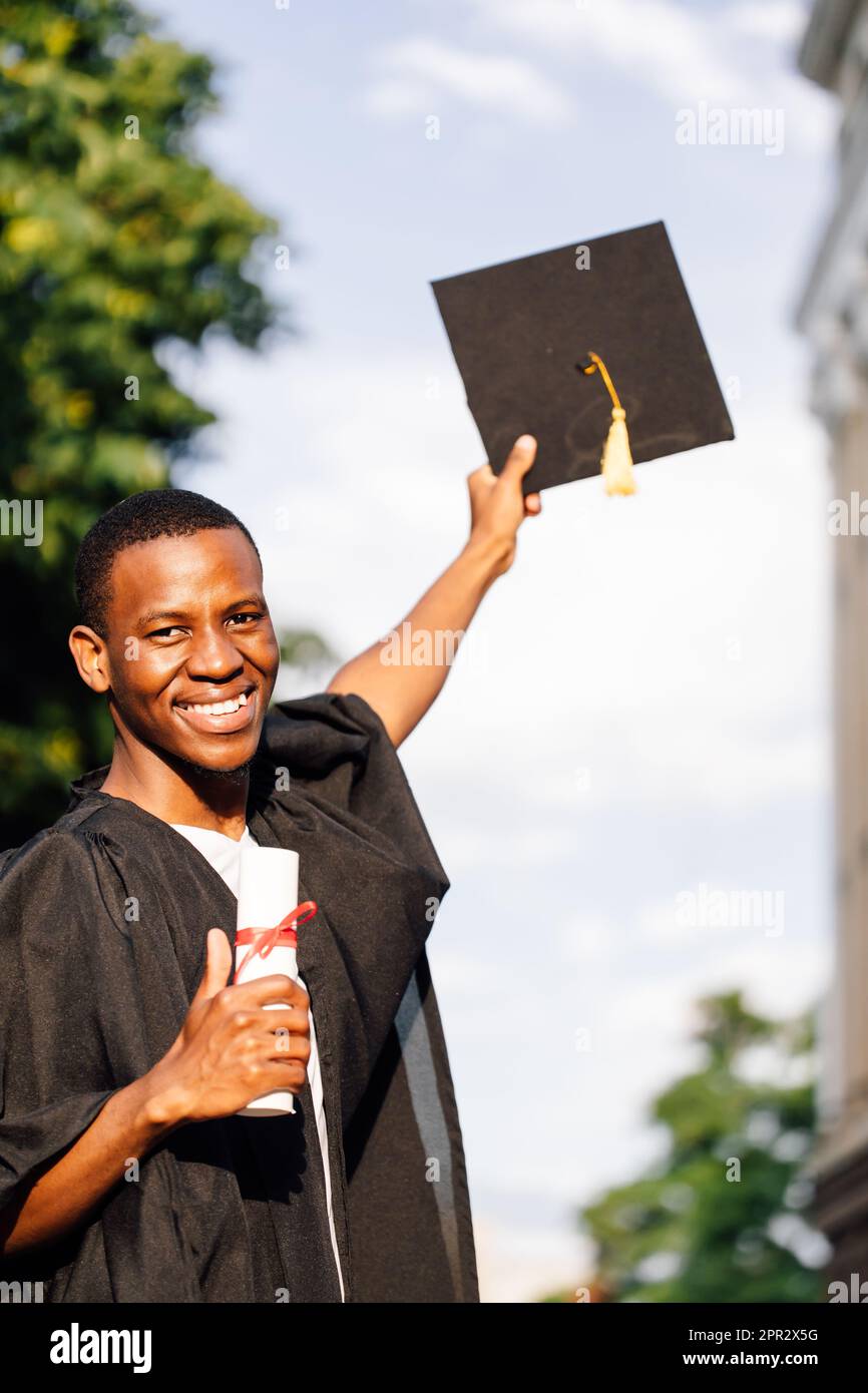 Joyeux diplômé afro-américain de l'université se tient dehors dans le  manteau et tient dans les mains diplôme d'études supérieures et chapeau  noir, ciel flou Photo Stock - Alamy