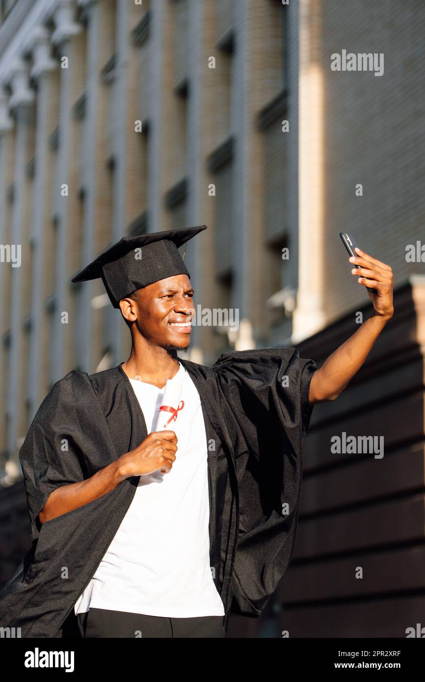 Un diplômé afro-américain souriant de l'université prend des photos avec l'appareil photo avant d'un smartphone avec un diplôme d'enseignement supérieur. Élève dans le manteau noir Banque D'Images