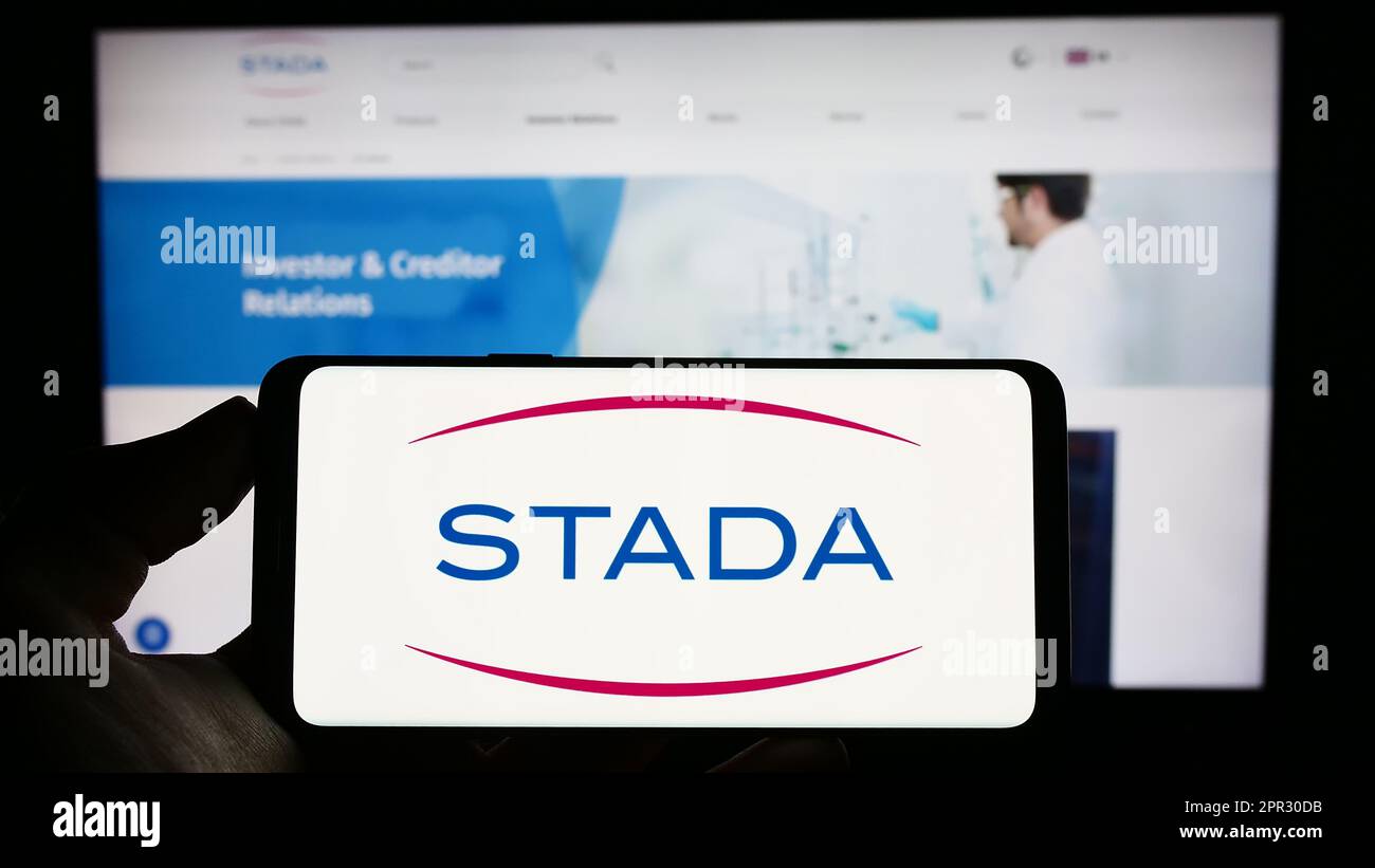 Personne tenant un téléphone portable avec le logo de la société allemande Stada Arzneimittel AG à l'écran en face de la page web d'affaires. Mise au point sur l'affichage du téléphone. Banque D'Images