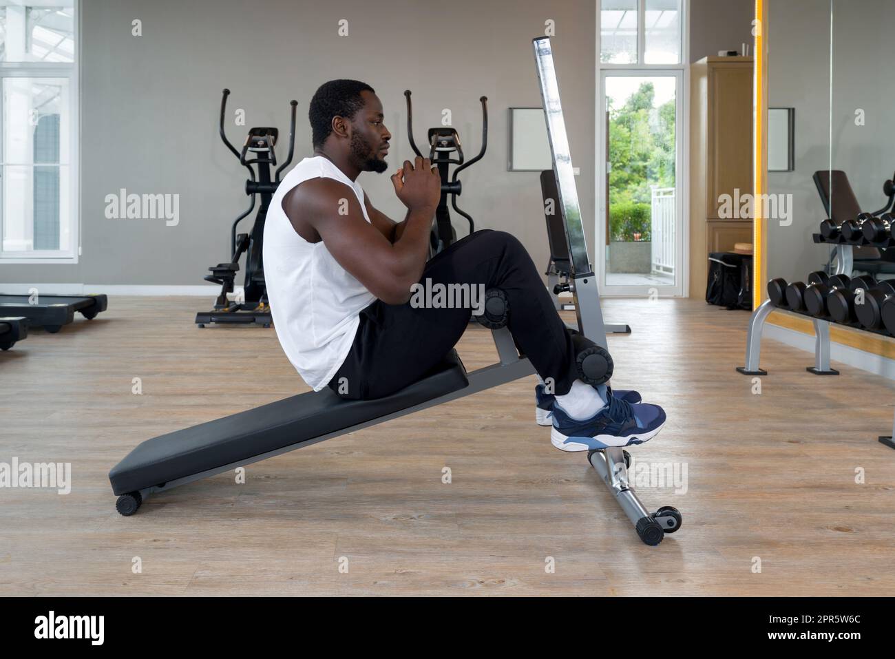 Jeune homme à cheveux courts et bouclés noir avec moustache et barbe assis  sur un banc multifonction. Il y a des appareils de cardio-training et un  jeu de haltères avec un rack