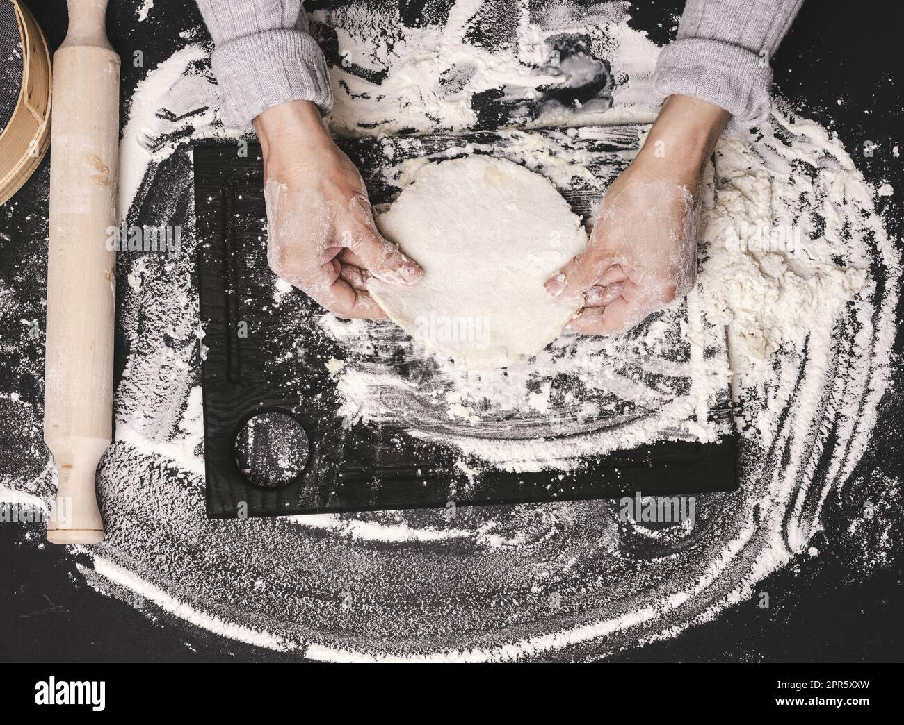 deux mains de femmes pétrient la pâte de farine de blé blanc sur une table noire, vue de dessus Banque D'Images