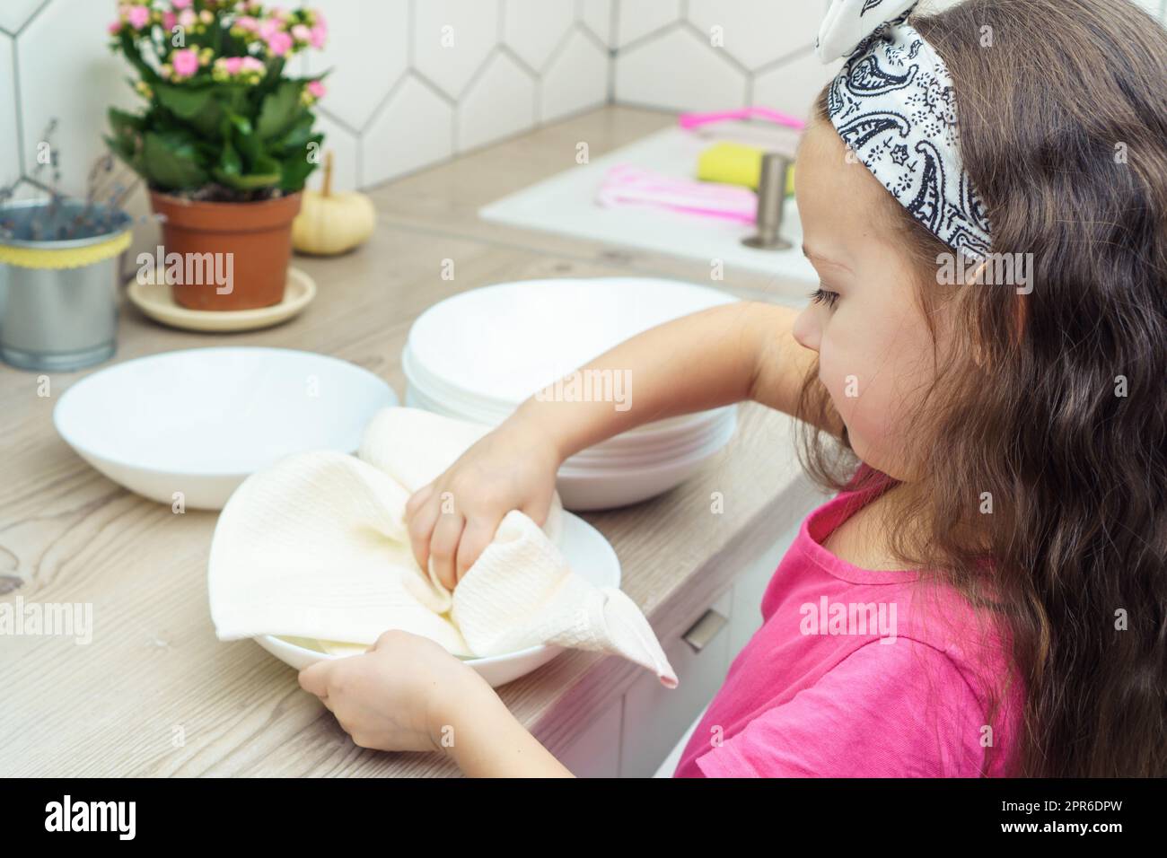 Vue latérale de la petite fille de préadolescence avec de longs cheveux foncés portant un T-shirt rose, laver la vaisselle, essuyer les assiettes propres avec un chiffon blanc près des fleurs sur la table Banque D'Images