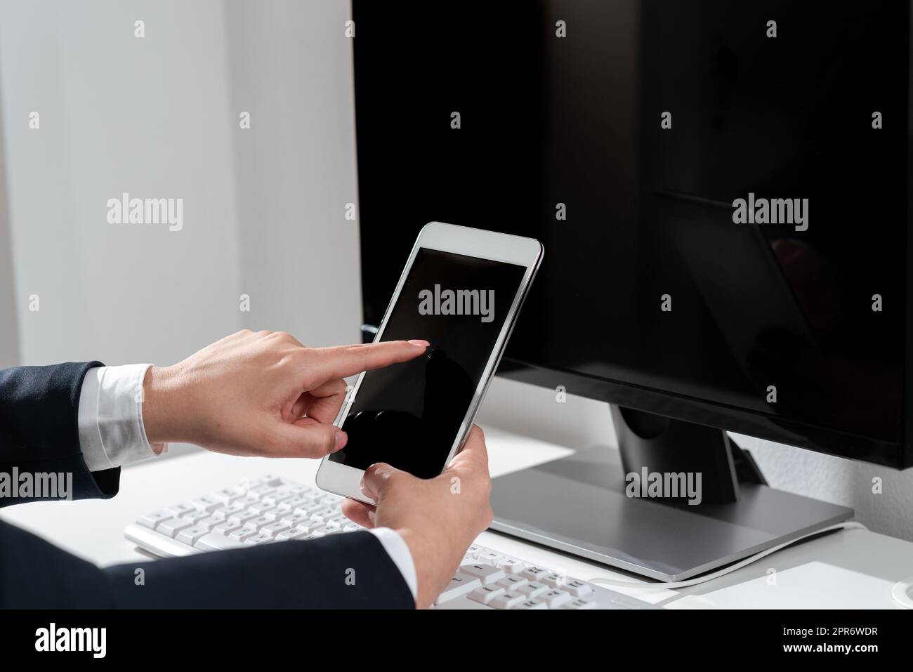 Assis sur le bureau avec ordinateur, femme d'affaires tenant le téléphone mobile et pointant avec un doigt dessus. Femme en costume présentant des informations crutiales d'une main. Banque D'Images