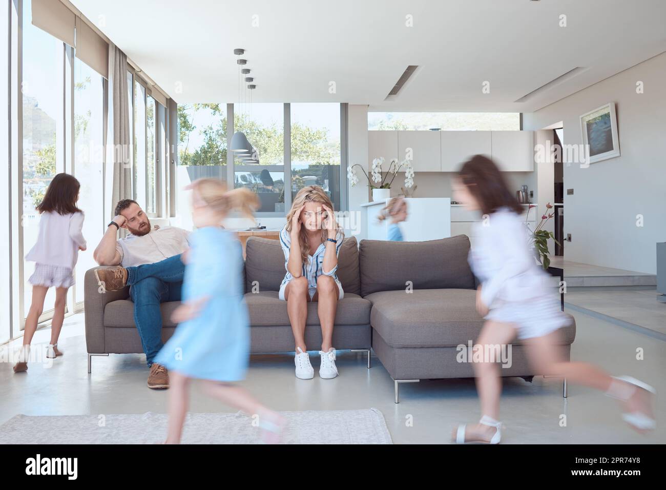 La mère et le père épuisés ont l'air stressés, tandis que les enfants jouent dans le salon. Bouleverser les parents avec des maux de tête assis sur le canapé, les enfants jouant autour d'eux. Jeunes filles jouant Banque D'Images