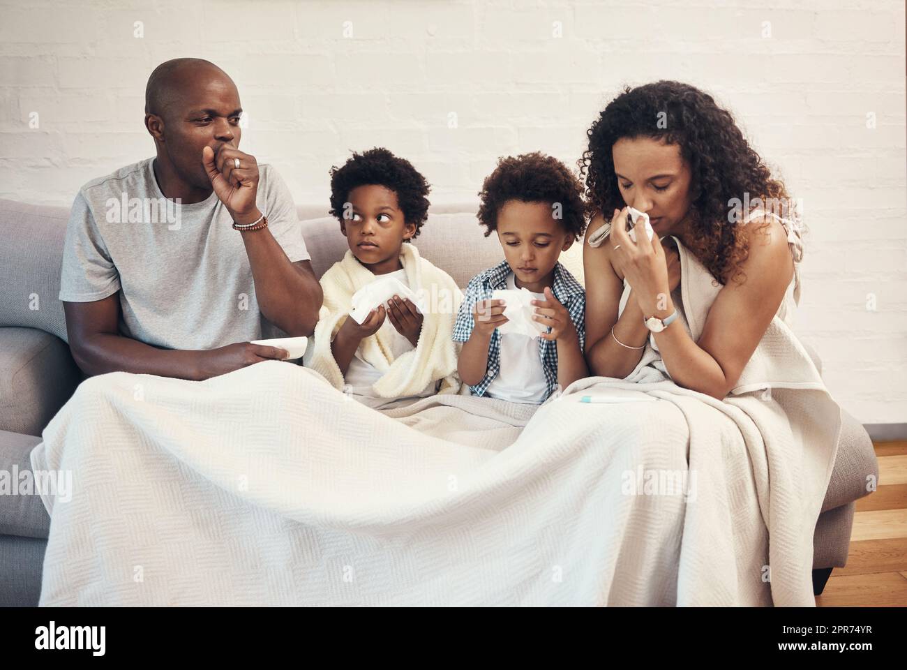 Famille afro-américaine de quatre personnes se sentant malade et malade avec la grippe et le froid alors qu'il est couvert dans une couverture sur un canapé. Les parents et deux enfants toussent et soufflent le nez à cause des symptômes d'allergie saisonniers et du covid Banque D'Images