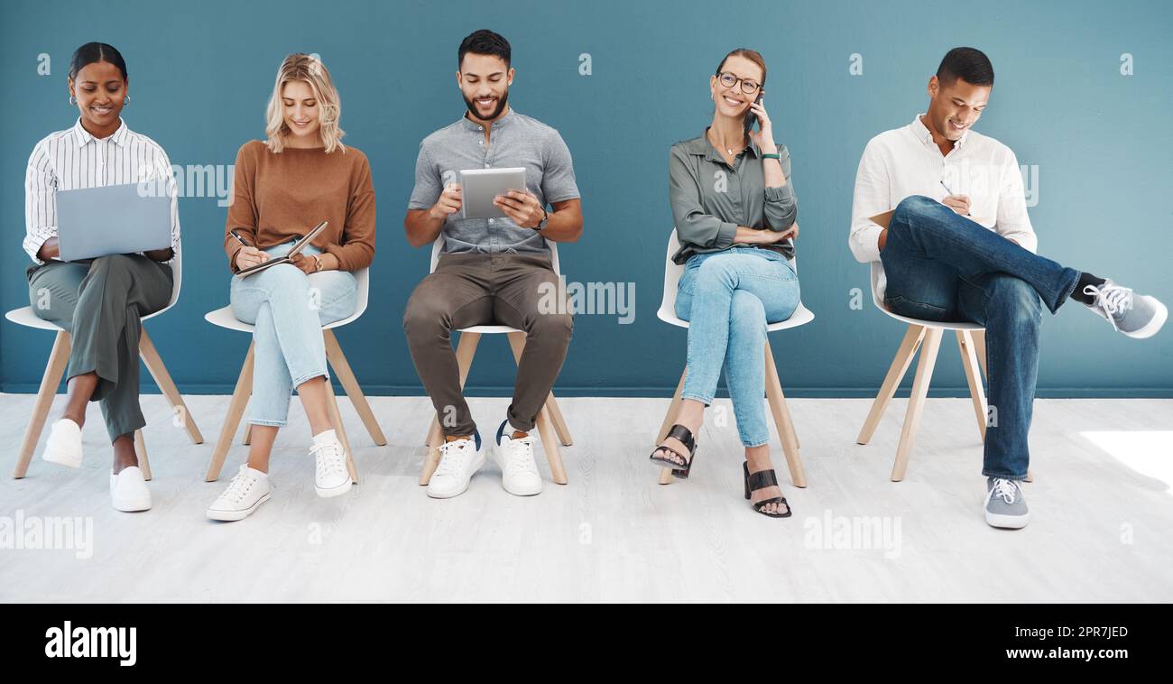 Un groupe diversifié d'hommes et de femmes d'affaires confiants qui naviguent sur des appareils technologiques tout en étant assis sur des chaises dans la salle d'attente sur un fond bleu. Les candidats et les candidats heureux et prometteurs sont prêts pour une entrevue de sélection en ligne pour une offre d'emploi Banque D'Images