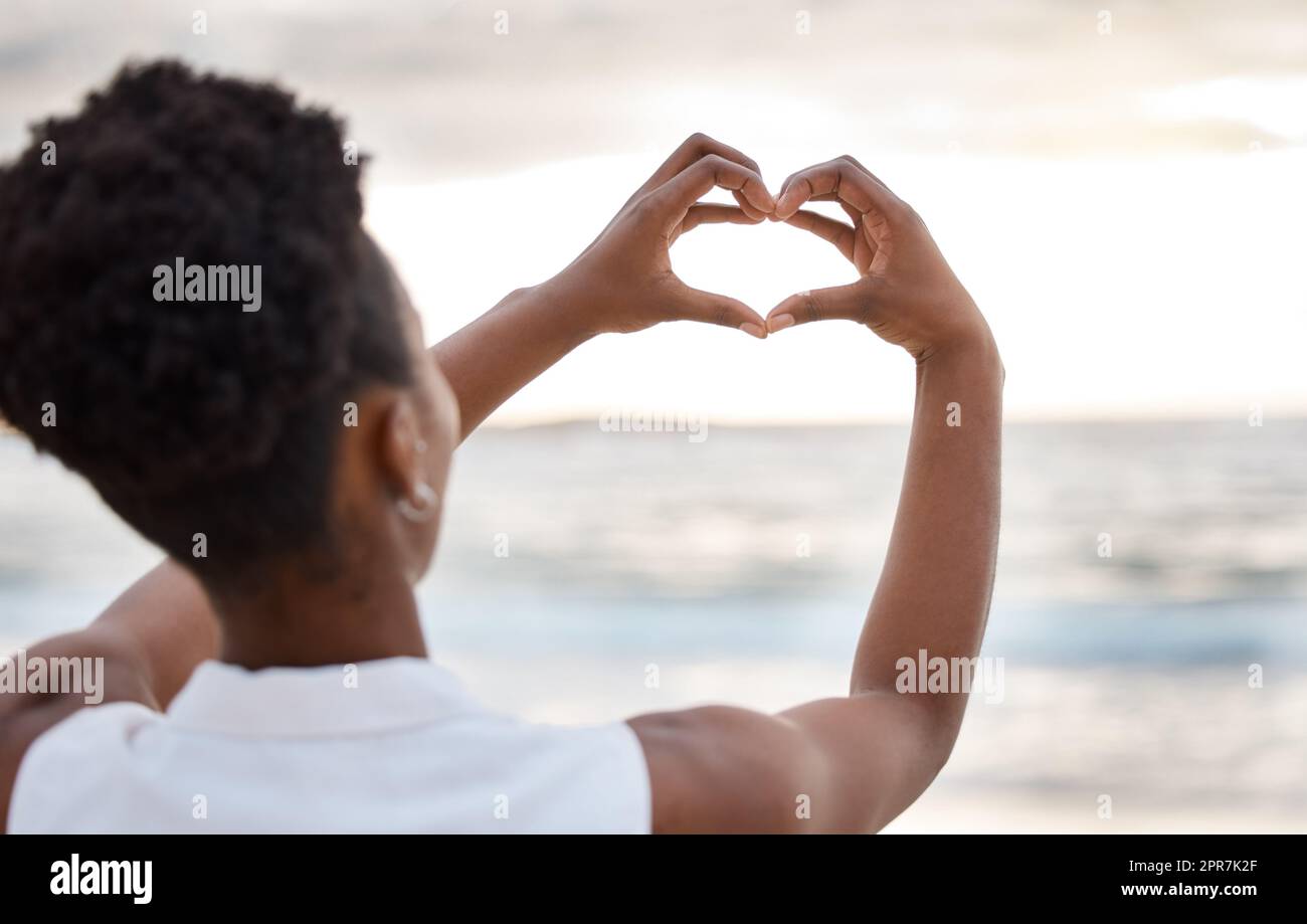 Vision arrière des mains sur une femme afro-américaine en train de faire un coeur contre le ciel à l'extérieur. Femme noire confiante debout sur une plage à l'extérieur au coucher du soleil se faisant un geste vers la mer. Santé et amour Banque D'Images