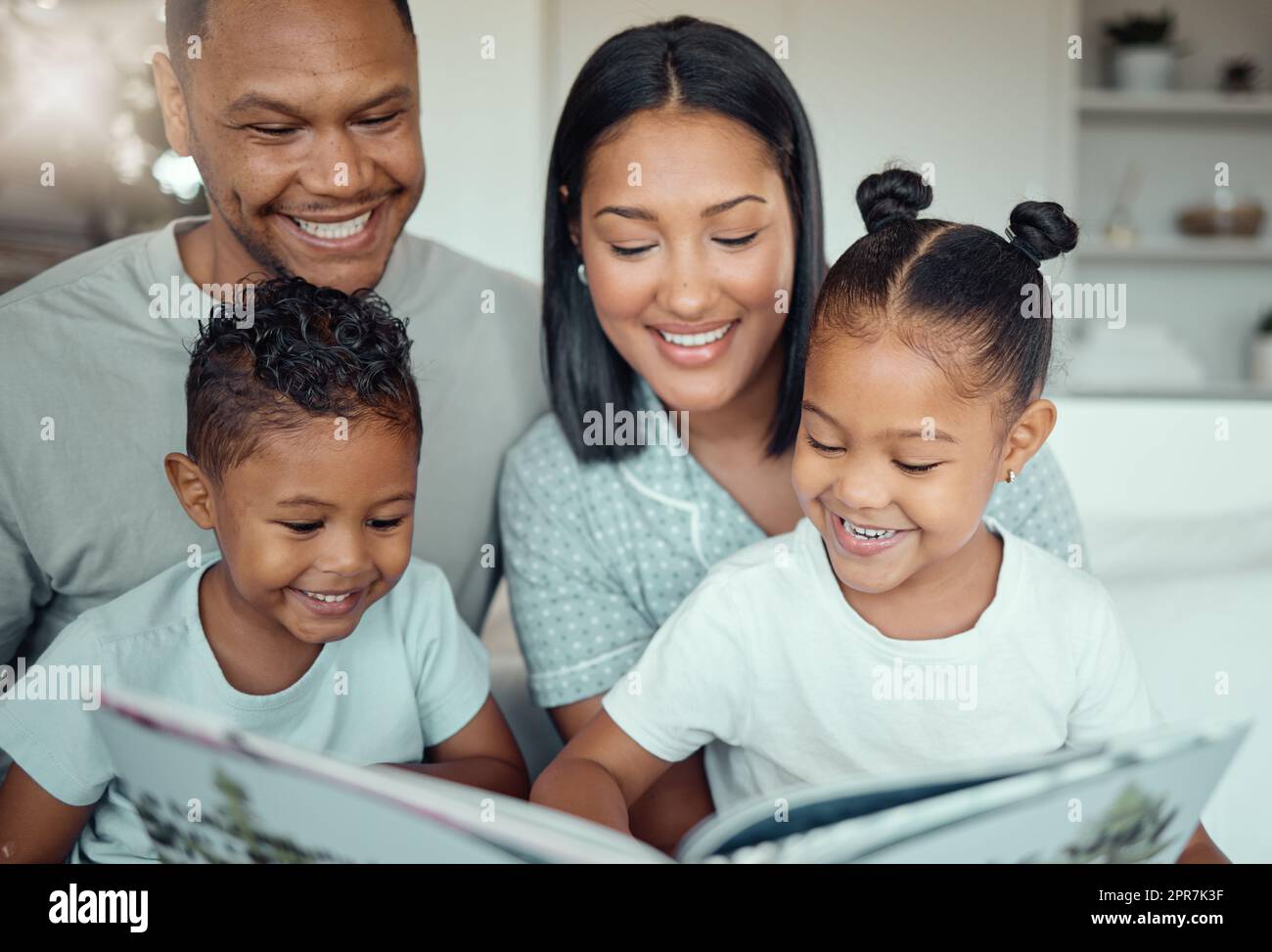 Les jeunes parents de race mixte maman et papa lisant un livre à petite fille et fils tout en liant et passer du temps ensemble à la maison le matin. Deux enfants qui apprécient l'histoire du conte de fées tout en étant assis avec leurs parents Banque D'Images