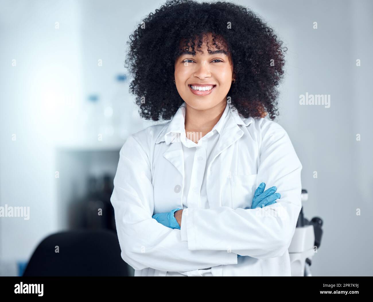 Portrait d'une belle jeune femme afro-américaine avec un afro portant un manteau et des gants tout en se tenant debout avec ses bras croisés dans le laboratoire. Une femme scientifique de race mixte souriant joyeusement Banque D'Images