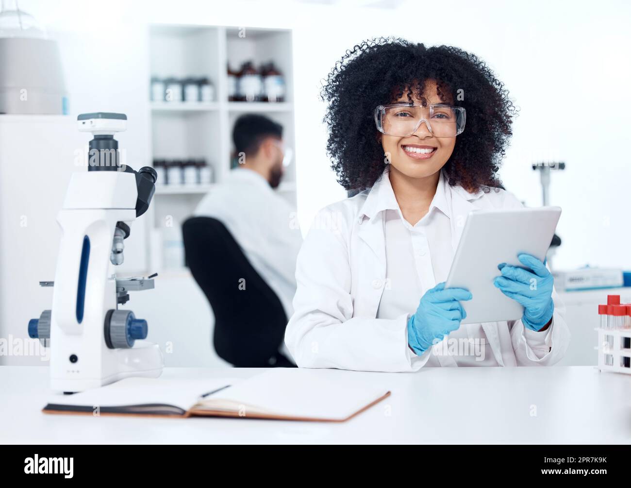 Portrait d'un jeune scientifique médical afro-américain heureux avec un afro utilisant une tablette numérique pour enregistrer les résultats de test à partir d'un microscope. Chimiste de race mixte découvrant une cure de vaccin dans un laboratoire Banque D'Images