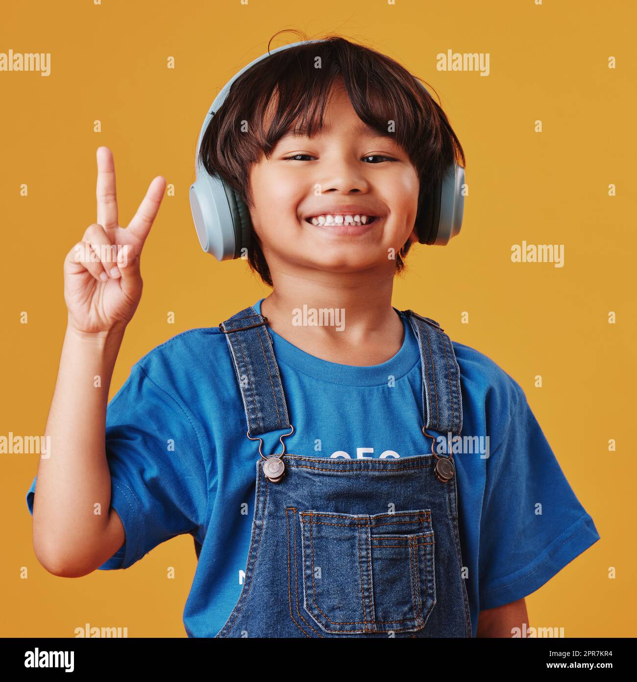 Un adorable petit garçon asiatique qui écoute de la musique tout en portant un casque et fait un geste de paix sur un fond d'espace publicitaire orange. Adorable enfant chinois qui ressent la magie de la musique Banque D'Images