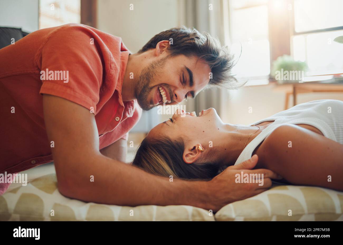 Bonne jeune femme couché tandis que son petit ami penche sur elle comme ils rient ensemble et regardent dans les autres yeux. Couple affectueux qui profite d'un moment romantique et intime chez soi Banque D'Images