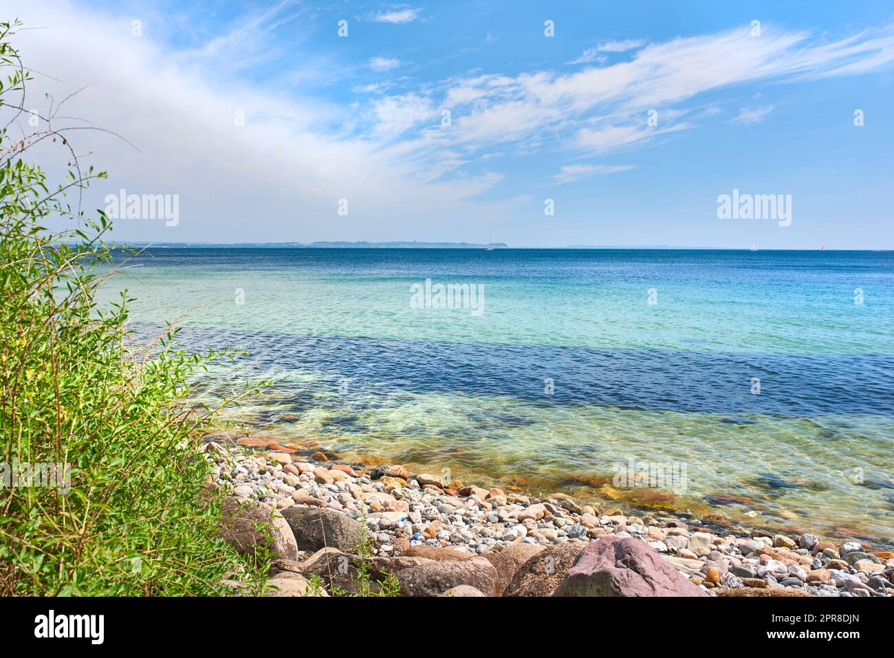 CopySpace en mer avec un ciel bleu ciel nuageux. L'océan calme se délave sur des pierres sur une plage vide avec des voiliers qui naviguent à l'horizon. Paysage pittoresque pour des vacances d'été relaxantes Banque D'Images