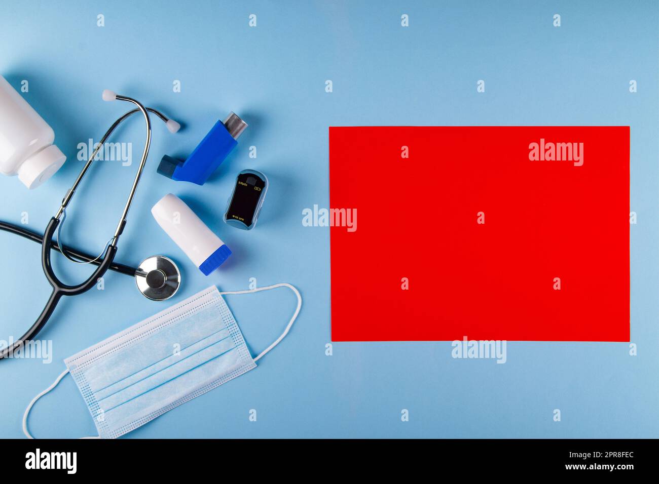 Vue de dessus du masque médical, stéthoscope, oxymètre, bouteille de pilules et inhalateurs sur fond bleu pastel avec feuille de papier rouge vif rectangulaire pour Banque D'Images