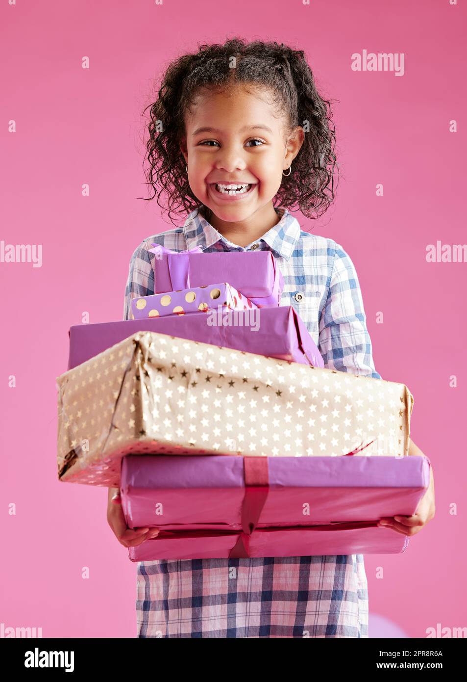 Une jolie petite fille de race mixte avec des cheveux bouclés tenant une pile de cadeaux enveloppés contre un arrière-plan rose copyspace dans un studio. Enfant africain à la recherche enthousiaste d'obtenir des cadeaux pour son anniversaire Banque D'Images