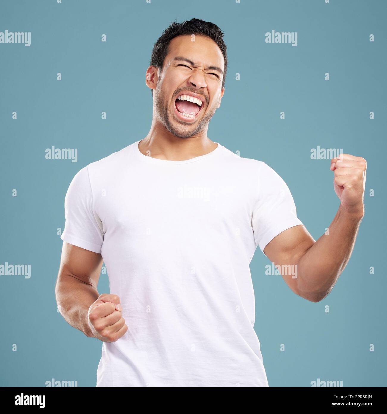 Beau jeune homme de course mixte célébrant la victoire ou le succès tout en se tenant en studio isolé sur fond bleu. Un homme hispanique applaudit et pompe ses poings au succès ou à la réussite Banque D'Images