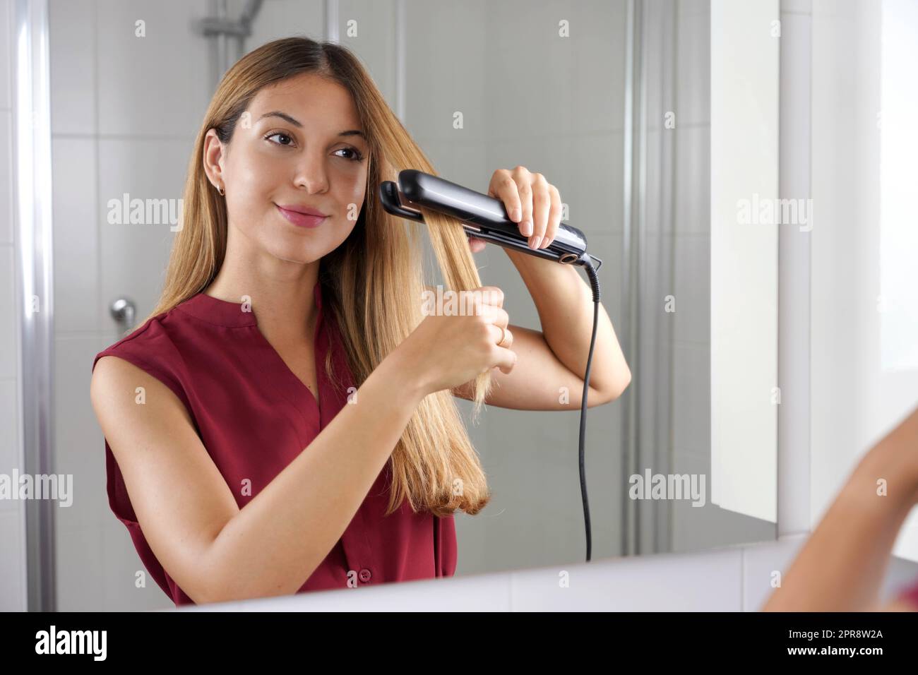 Jeune femme hispanique utilisant un lisseur à vapeur pour coiffer les cheveux dans le miroir de la salle de bains Banque D'Images