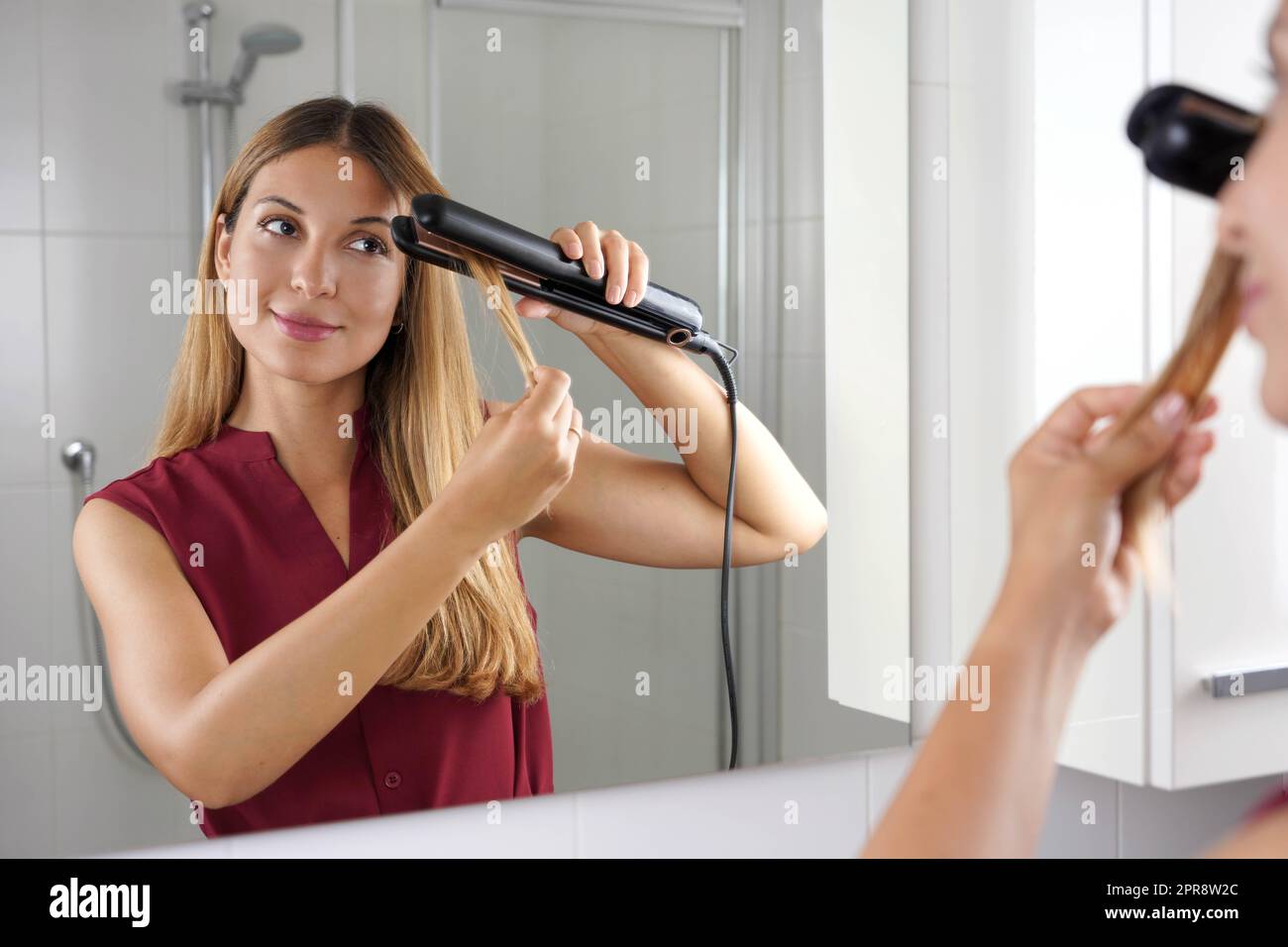 Fille utilisant un lisseur vapeur pour coiffer les cheveux au miroir de la salle de bains Banque D'Images