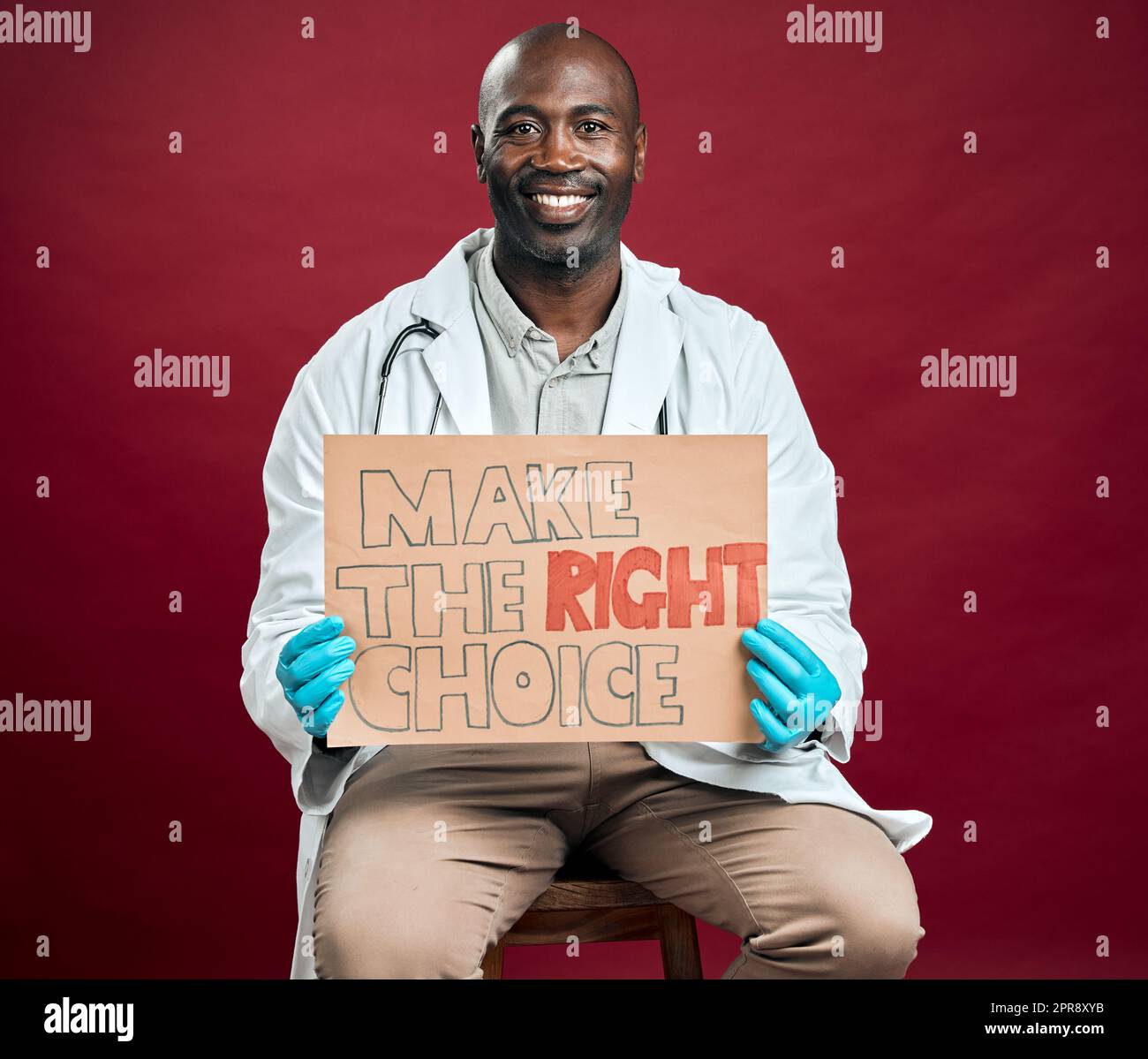Un médecin afro-américain covid tient et montre une affiche. Portrait d'un médecin noir souriant isolé sur fond rouge studio avec copyspace. Homme faisant la promotion et encourageant le vaccin corona en signe Banque D'Images
