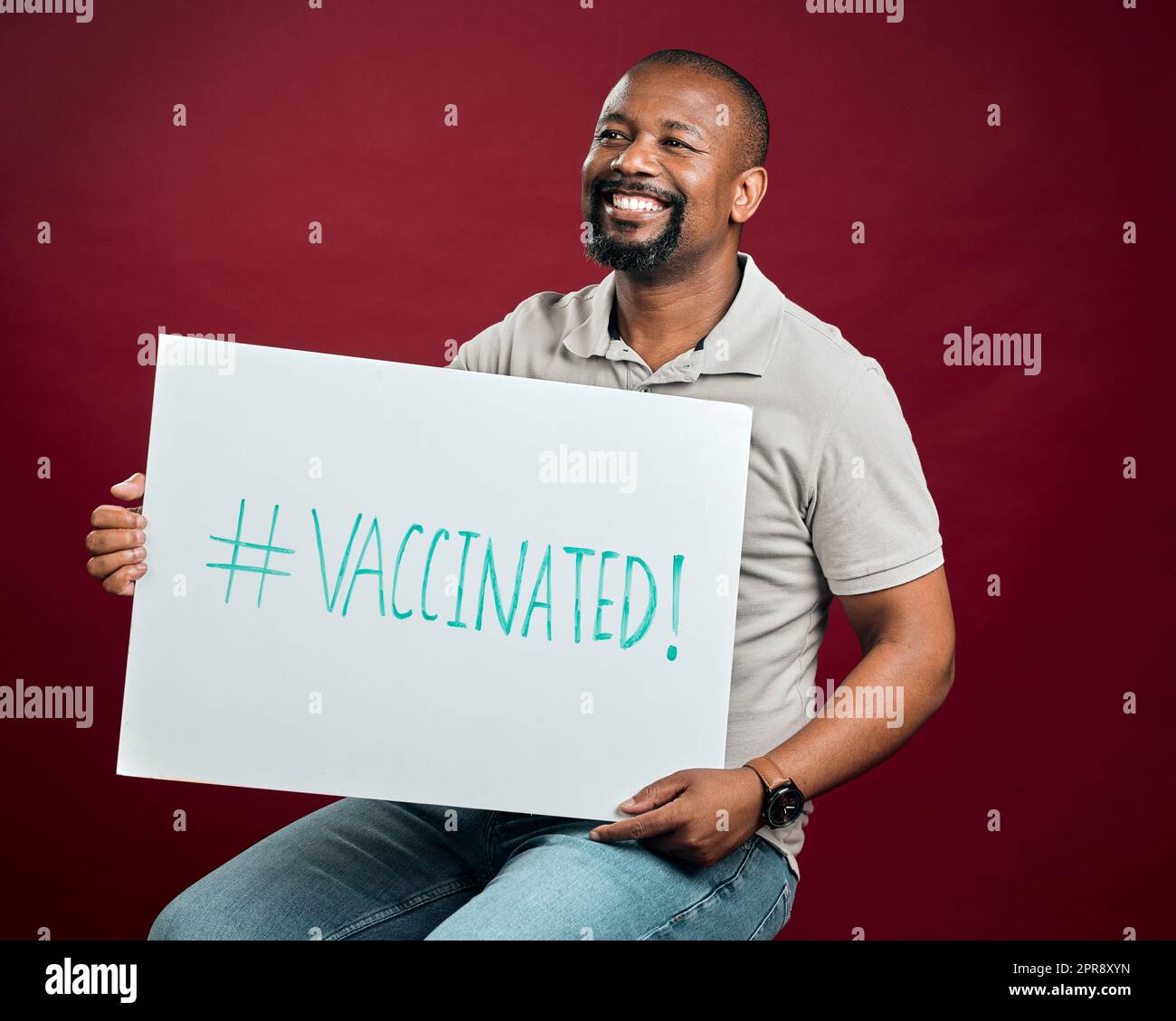 Homme vacciné covid afro-américain montrant et tenant une affiche. Homme noir souriant isolé contre arrière-plan rouge studio avec espace de copie. Modèle heureux utilisant le signe pour promouvoir le vaccin corona et motiver Banque D'Images