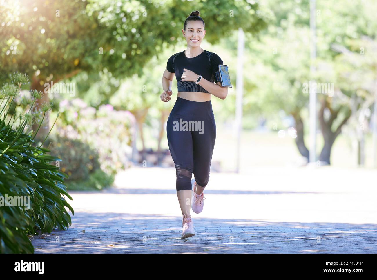 Bonne sportive jeune athlète féminine portant un brassard de téléphone et une montre intelligente tout en faisant du jogging au parc par une journée ensoleillée. Jeune femme hispanique qui fait de l'exercice à l'extérieur dans la nature Banque D'Images