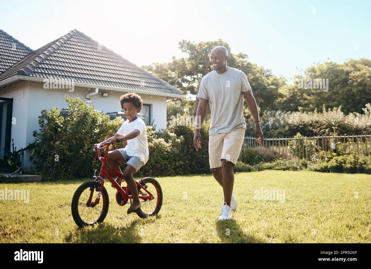 Adorable petit garçon afro-américain apprenant à faire son vélo dehors avec son père. Papa et fils s'amusent dans leur arrière-cour lors d'une journée ensoleillée Banque D'Images