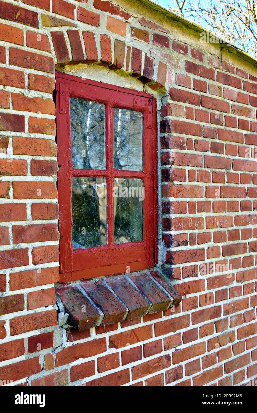 Vieille fenêtre sale dans une maison en briques rouges. Ancienne casaque avec cadre en bois rouge dans un bâtiment historique à la texture de peinture bosselée. Détails extérieurs d'un seuil de fenêtre dans une ville ou un village traditionnel Banque D'Images