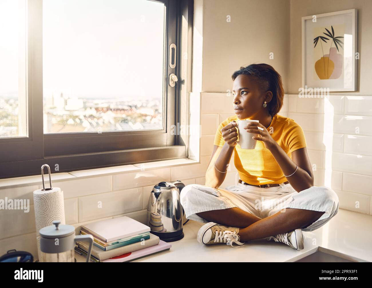 Pensant, femme sérieuse et africaine de boire du café tout en étant assis sur le comptoir de cuisine seul à la maison. Une femme noire, insouciante et paisible, savourant une tasse de thé tout en ayant l'air attentionnés Banque D'Images