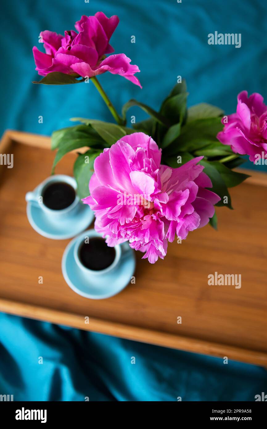 Belles pivoines rose dans un vase de verre et deux tasses de café sont debout sur un plateau en bois dans le lit. Banque D'Images