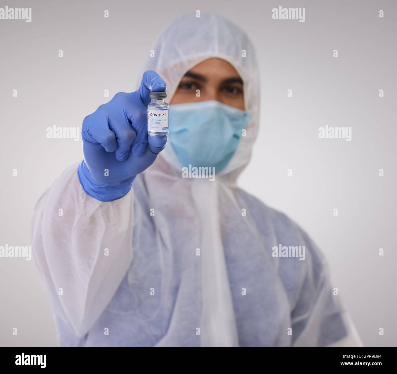 Croyez-moi, vous avez besoin de cela. Un professionnel de la santé debout dans un costume de noisette jetable et tenant un flacon pour le vaccin Covid. Banque D'Images