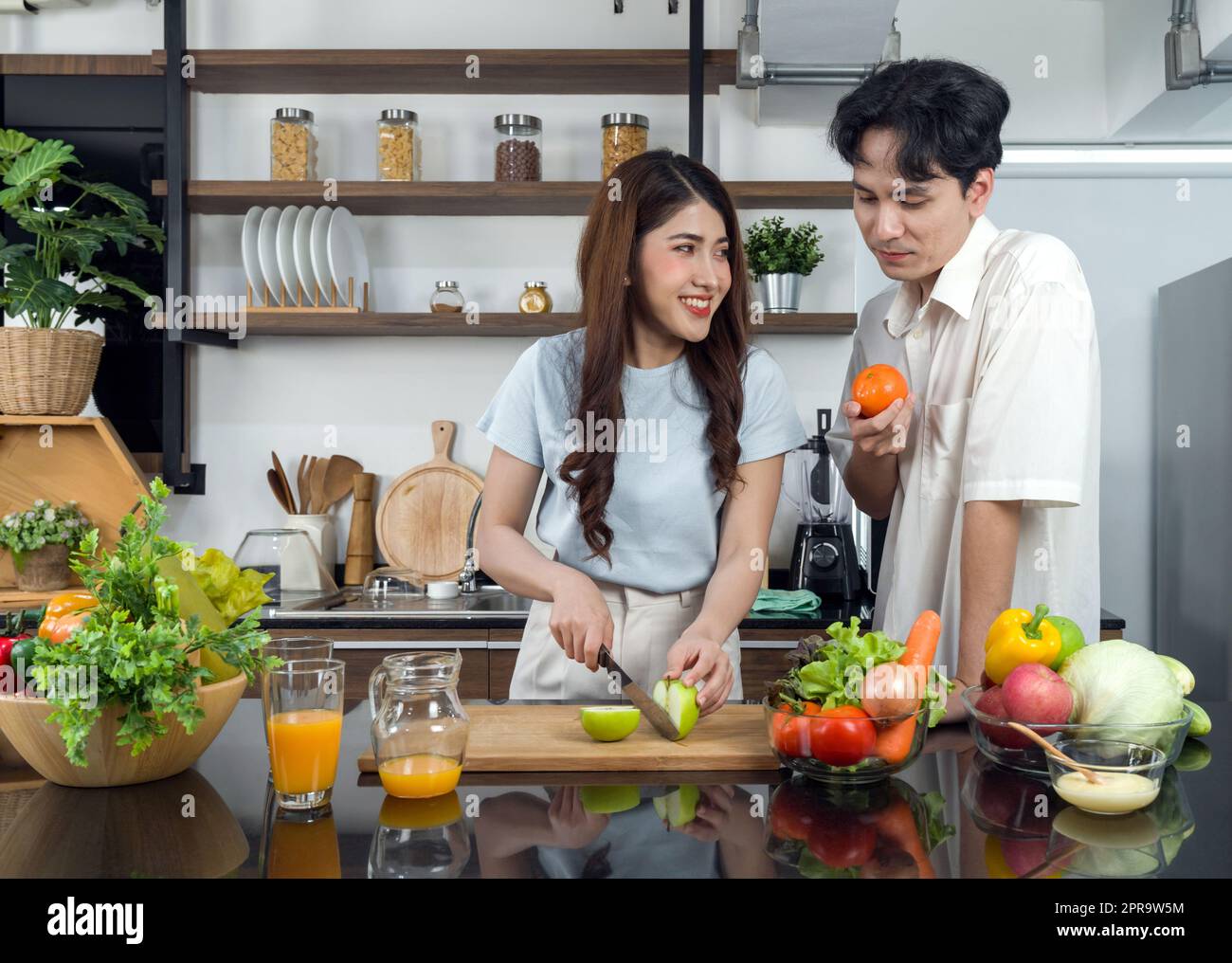 Un couple asiatique passe du temps ensemble dans la cuisine. Une jeune femme coupe une pomme verte sur une planche à découper en bois tandis que son petit ami se tient à côté d'elle avec une orange dans la main. Le jus de fruits est sur le comptoir. Banque D'Images